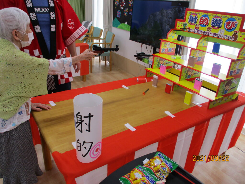 高齢者が参加する射的ゲーム。赤と白の装飾された屋台で季節イベントを開催中。
