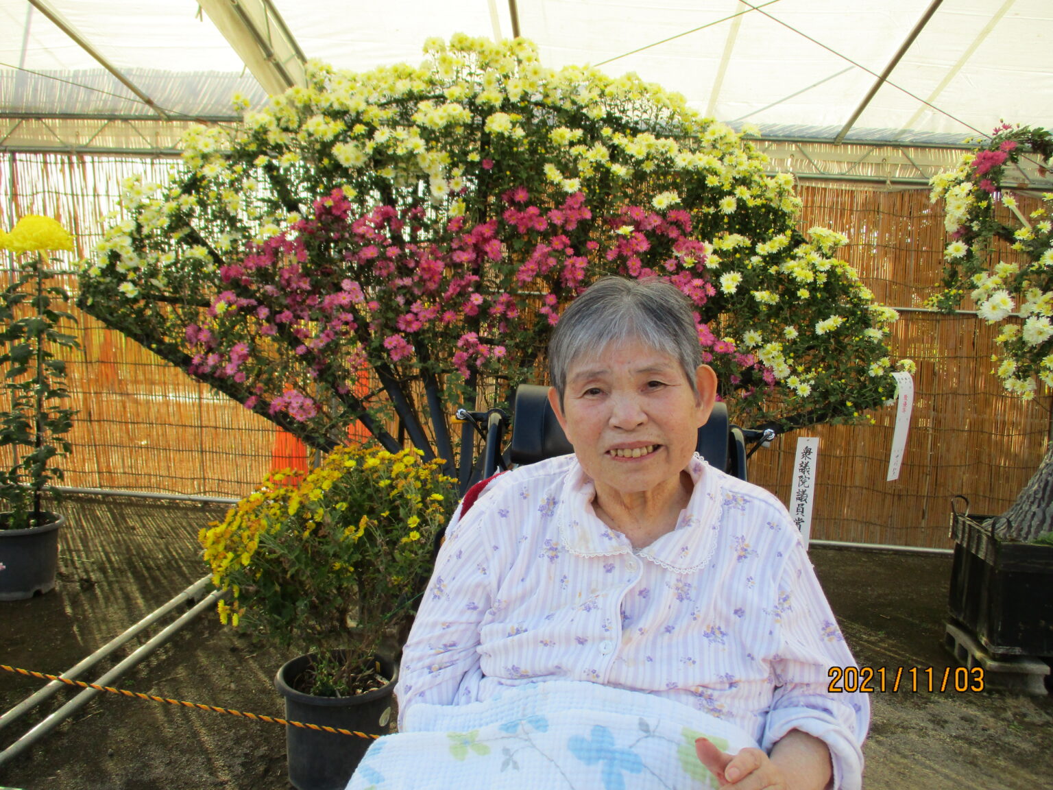 温室内の花々に囲まれた入居者の笑顔。園芸活動・レクリエーションの様子。